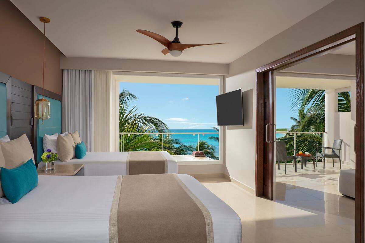 A hotel room with a bed, a TV, a ceiling fan, and a balcony with a view of the ocean.