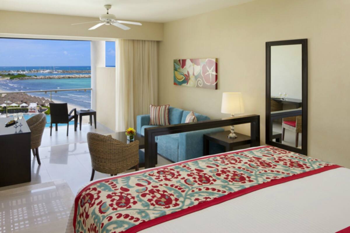 A hotel room with a large bed, a desk, a chair, a sofa, a table with a lamp, a painting, and a view of the ocean.