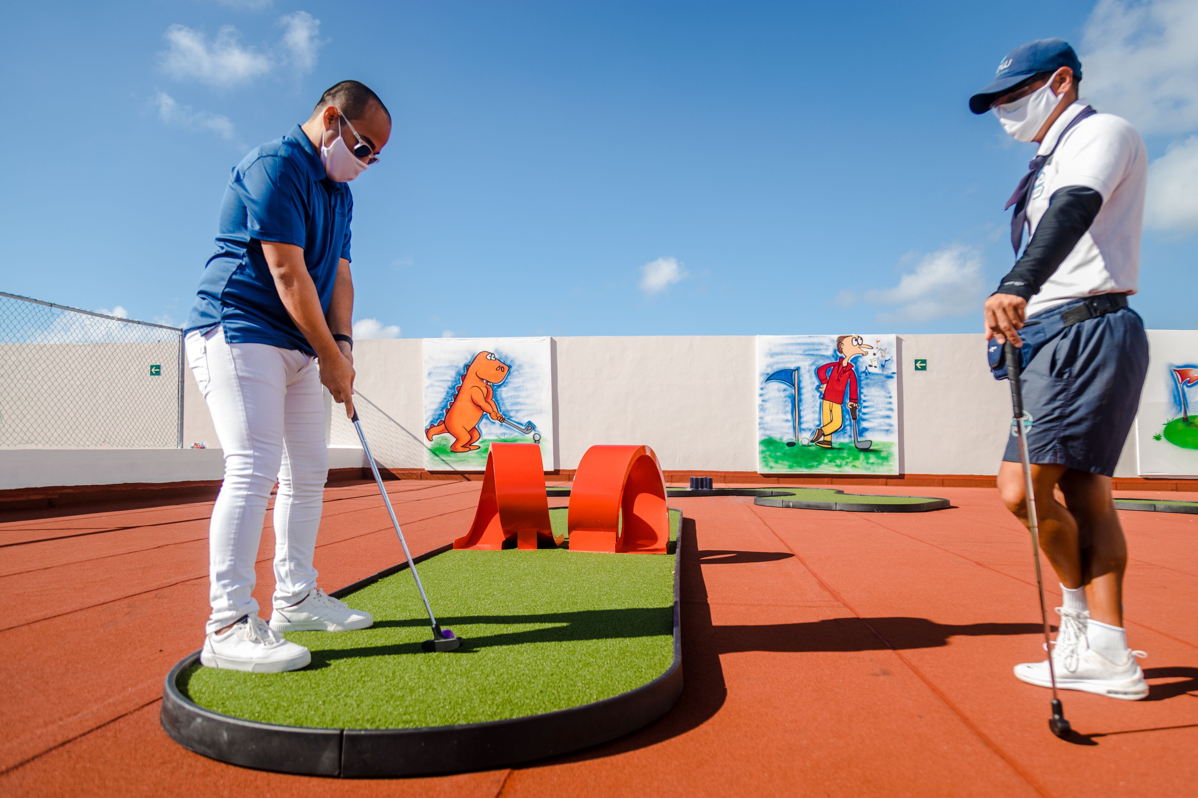 Un camp de mini golf amb una zona de posició verda, obstacles taronja i murals de dibuixos animats a les parets.