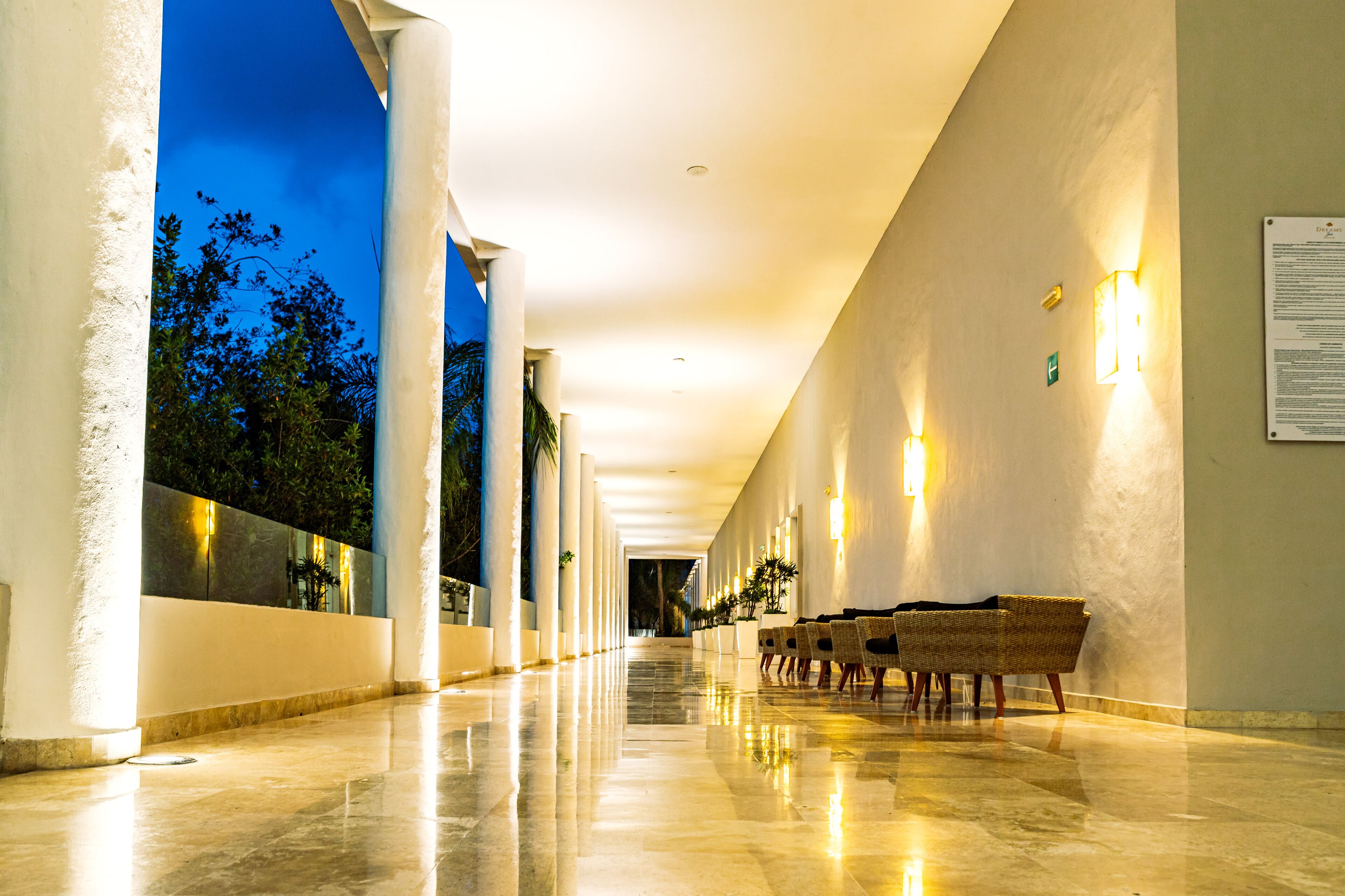 A corridor with a polished floor, white columns, and wicker chairs along one side.
