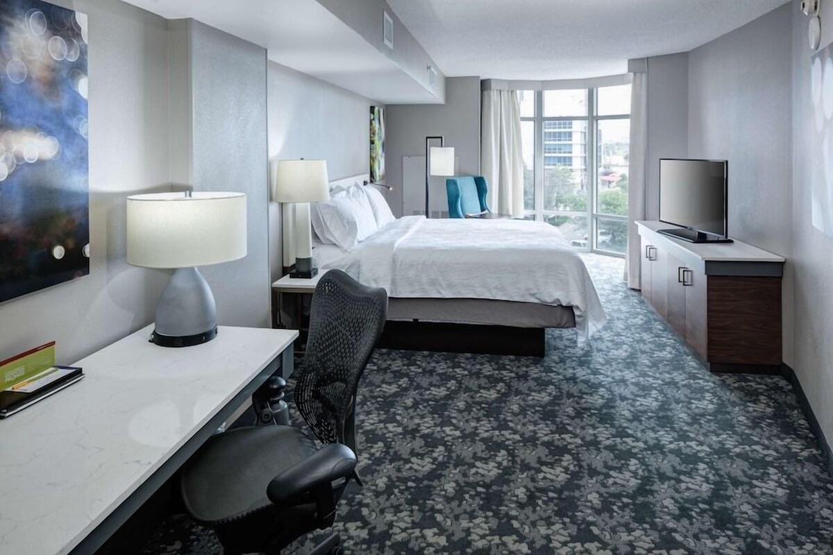 Une chambre d’hôtel équipée d’un lit, d’un bureau, d’une chaise, d’une télévision et d’une grande fenêtre donnant sur la ville.