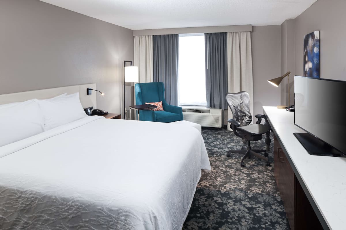 Ein Hotelzimmer mit einem großen Bett, einem Schreibtisch mit Fernseher, einem Sessel und einem Fenster mit Vorhängen.