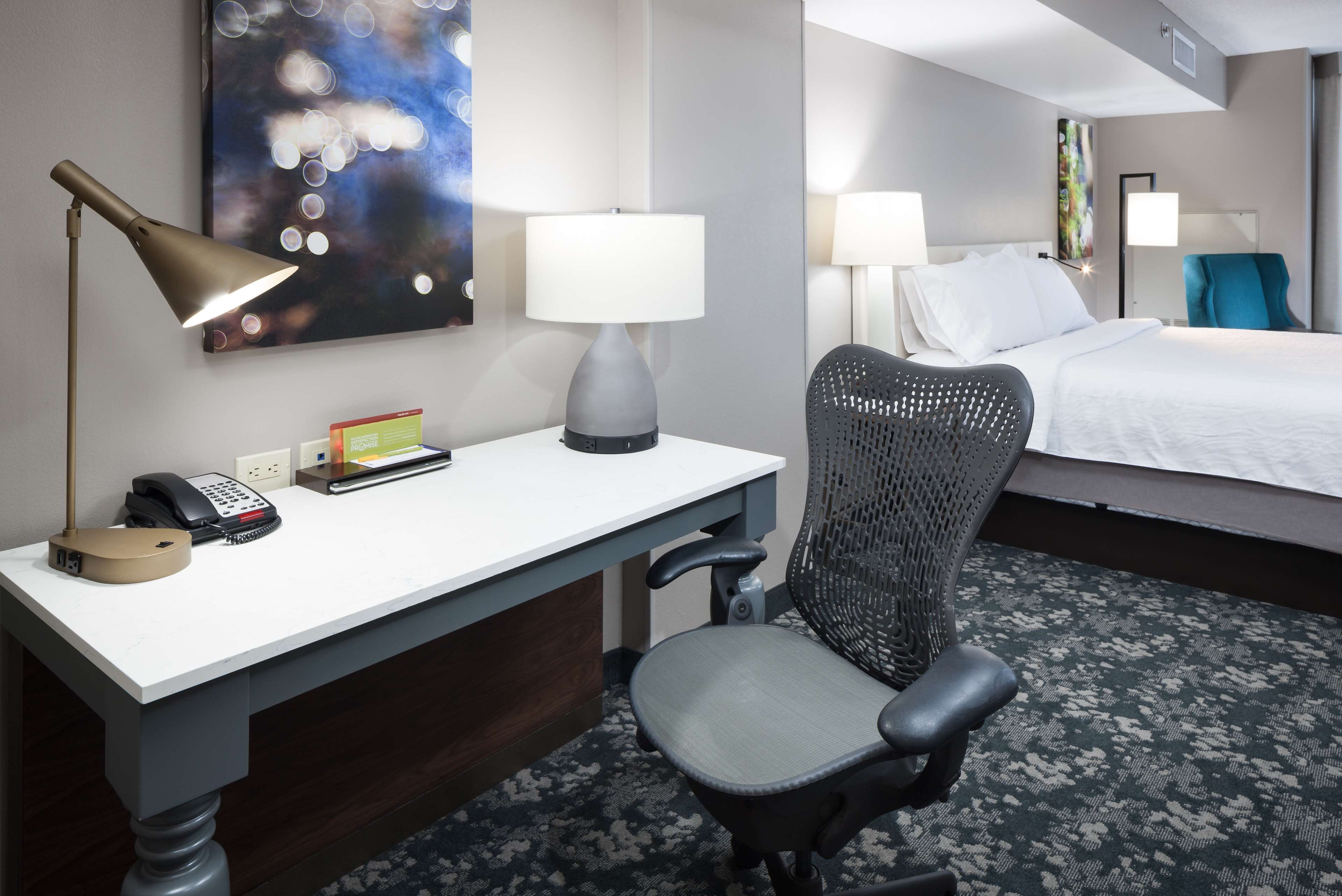 A hotel room with a desk, chair, bed, lamps, and artwork.