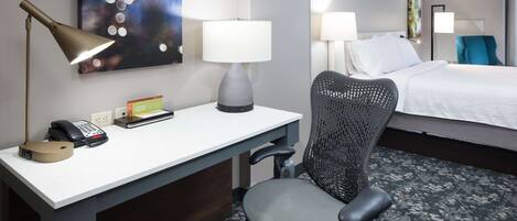 Habitación de hotel con escritorio, silla, cama, lámparas y obras de arte.