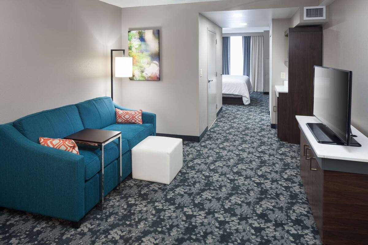 Ein Hotelzimmer mit einem blauen Sofa, einem kleinen weißen Hocker, einem Flachbildfernseher, einem Schreibtisch und einem Bild an der Wand.