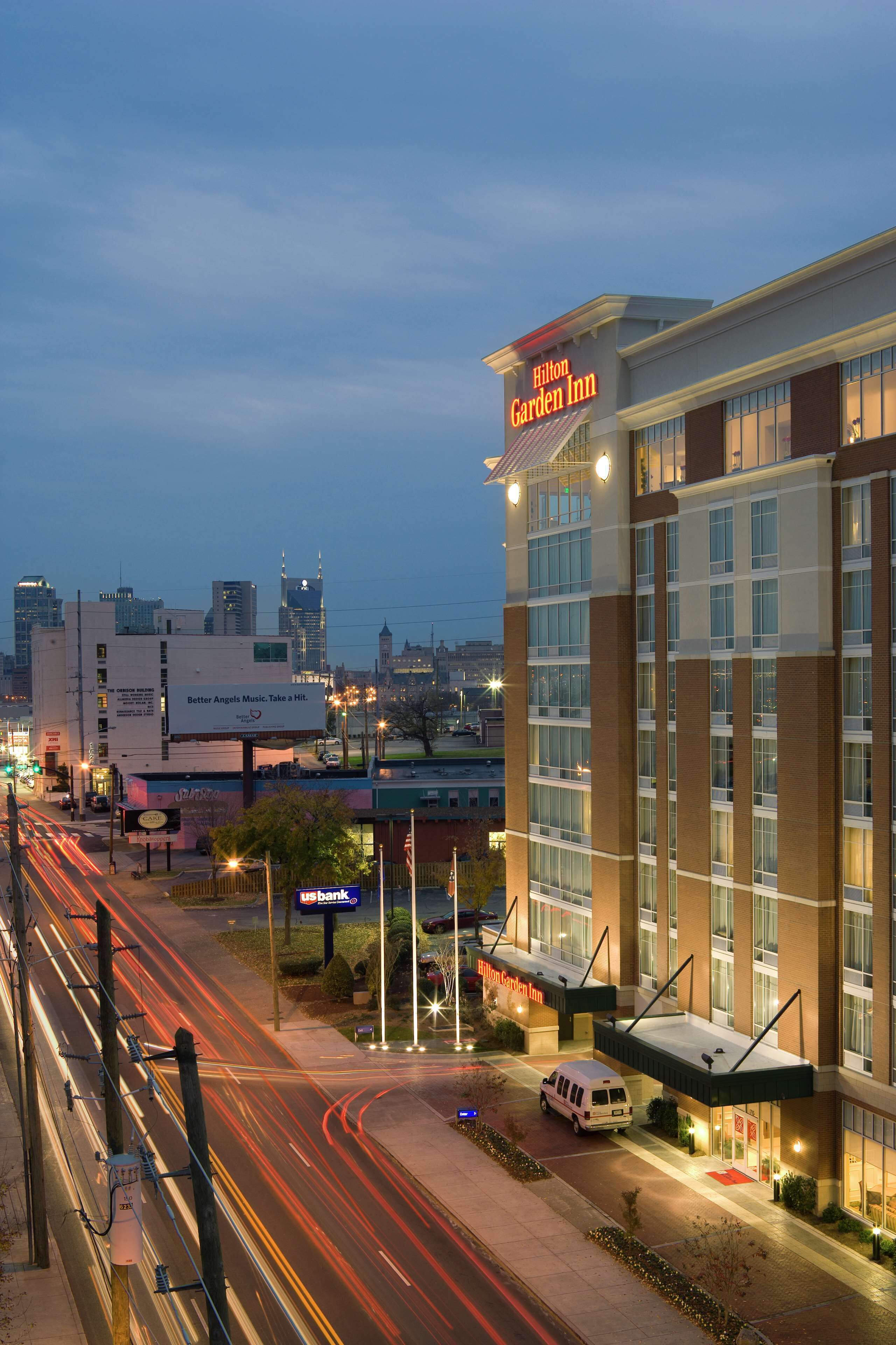 Un Hilton Garden Inn dal design moderno, situato vicino a una strada trafficata con semafori e lampioni ben visibili.