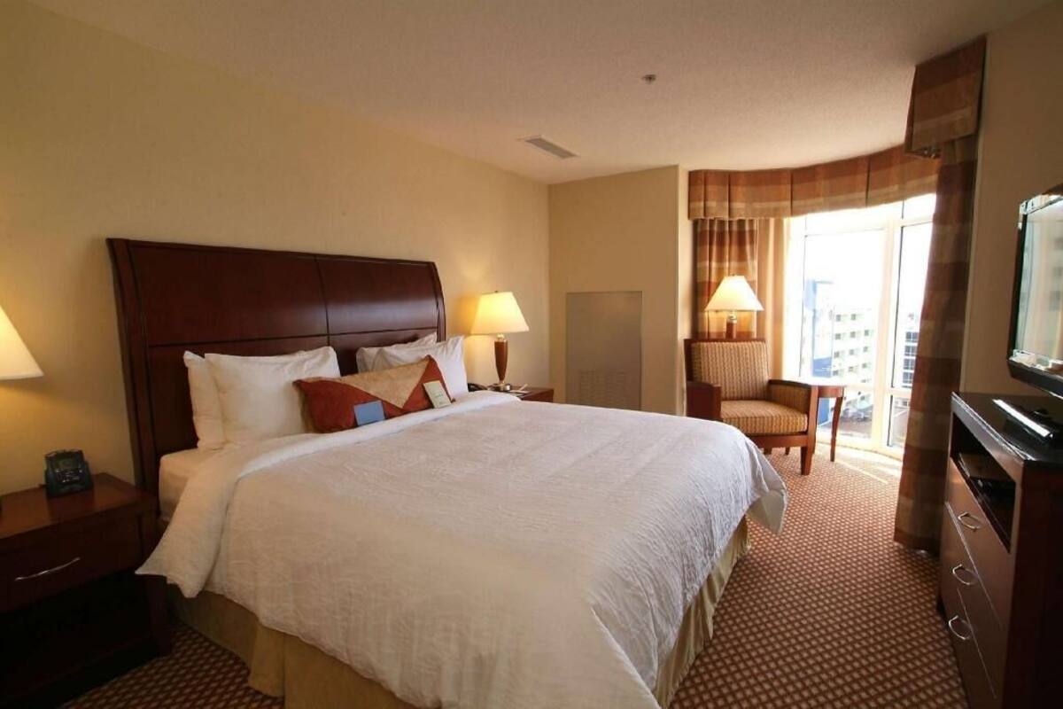 Ein Hotelzimmer mit einem großen Bett, zwei Nachttischen mit Lampen, einem Sessel, einem Schreibtisch mit Fernseher und einem großen Fenster mit Vorhängen.
