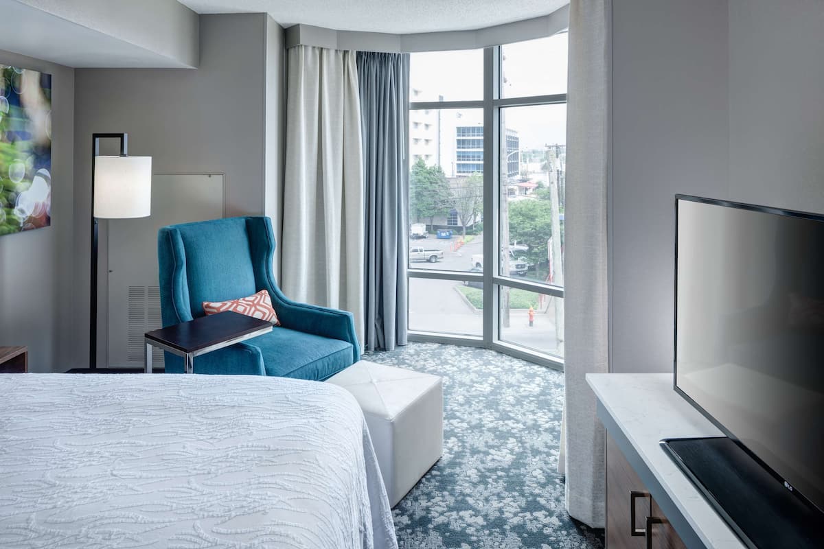 Ein Hotelzimmer mit einem großen Bett, einem blauen Sessel, einem Flachbildfernseher und einem großen Fenster mit Blick auf die Stadt.