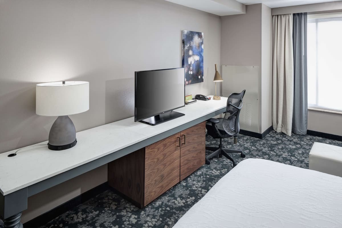 Ein Hotelzimmer mit Bett, Schreibtisch mit Computer, Lampe, Stuhl und einem Bild an der Wand.