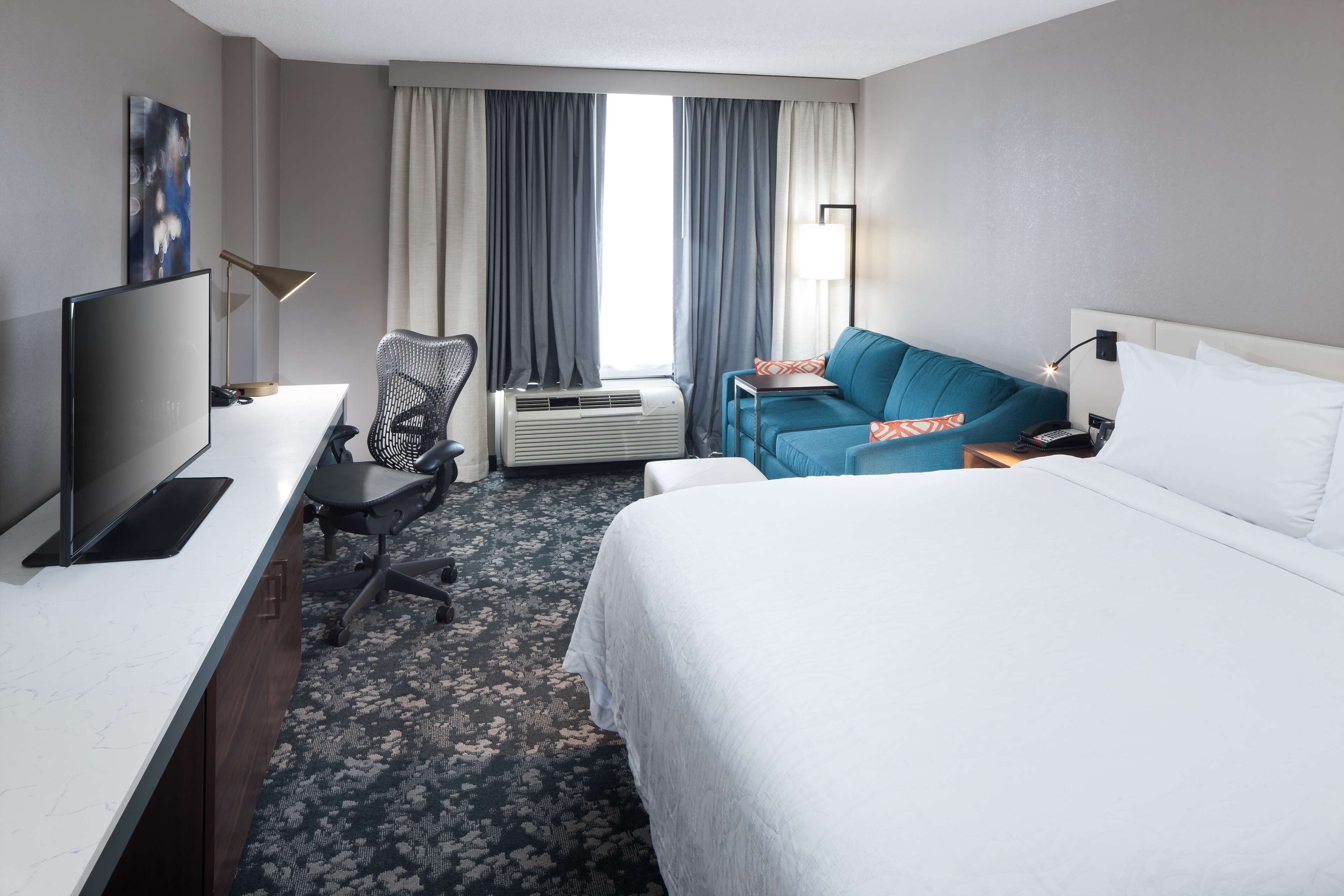 Habitación de hotel con cama, escritorio con televisor, una silla, sofá y ventana con cortinas.