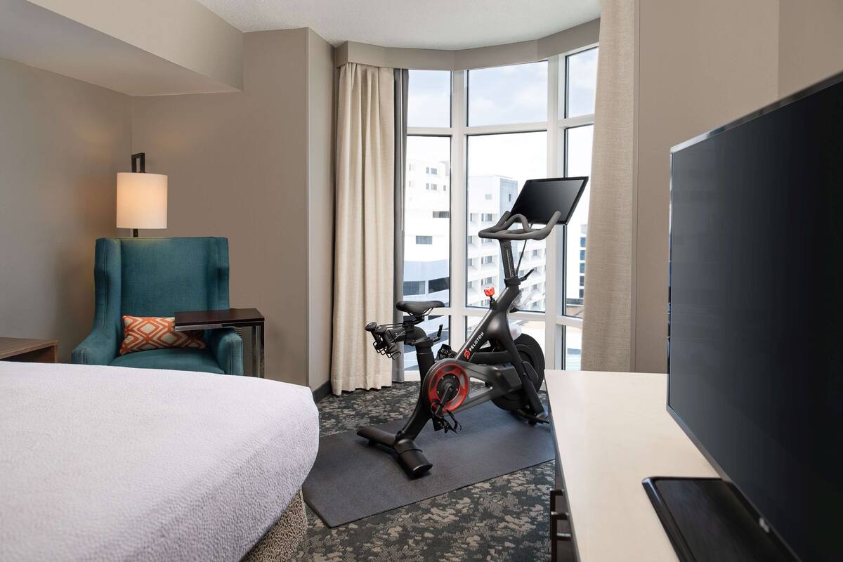 Ein Hotelzimmer mit einem Bett, einem Schreibtisch mit Computer, einem Crosstrainer, einem Fernseher und einem Fenster mit Blick auf Gebäude.