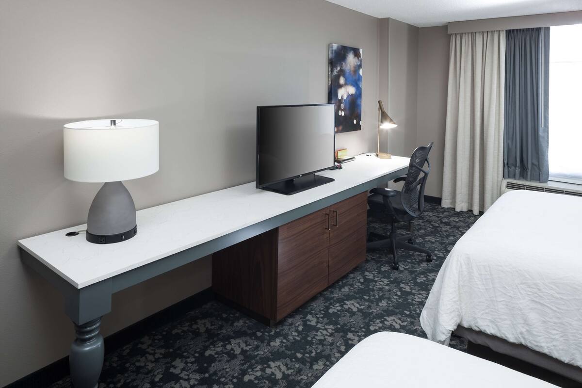 Ein Hotelzimmer mit einem Bett, einem Schreibtisch mit Fernseher, einer Lampe, einem Sessel und einem Bild an der Wand.