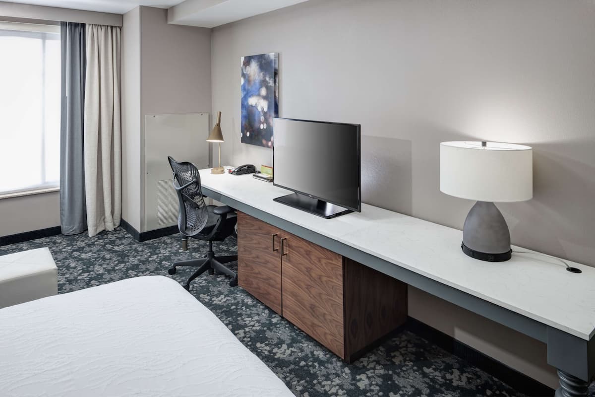 Ein Hotelzimmer mit Bett, Schreibtisch mit Computer, Lampe, Stuhl und einem Fenster mit Vorhängen.