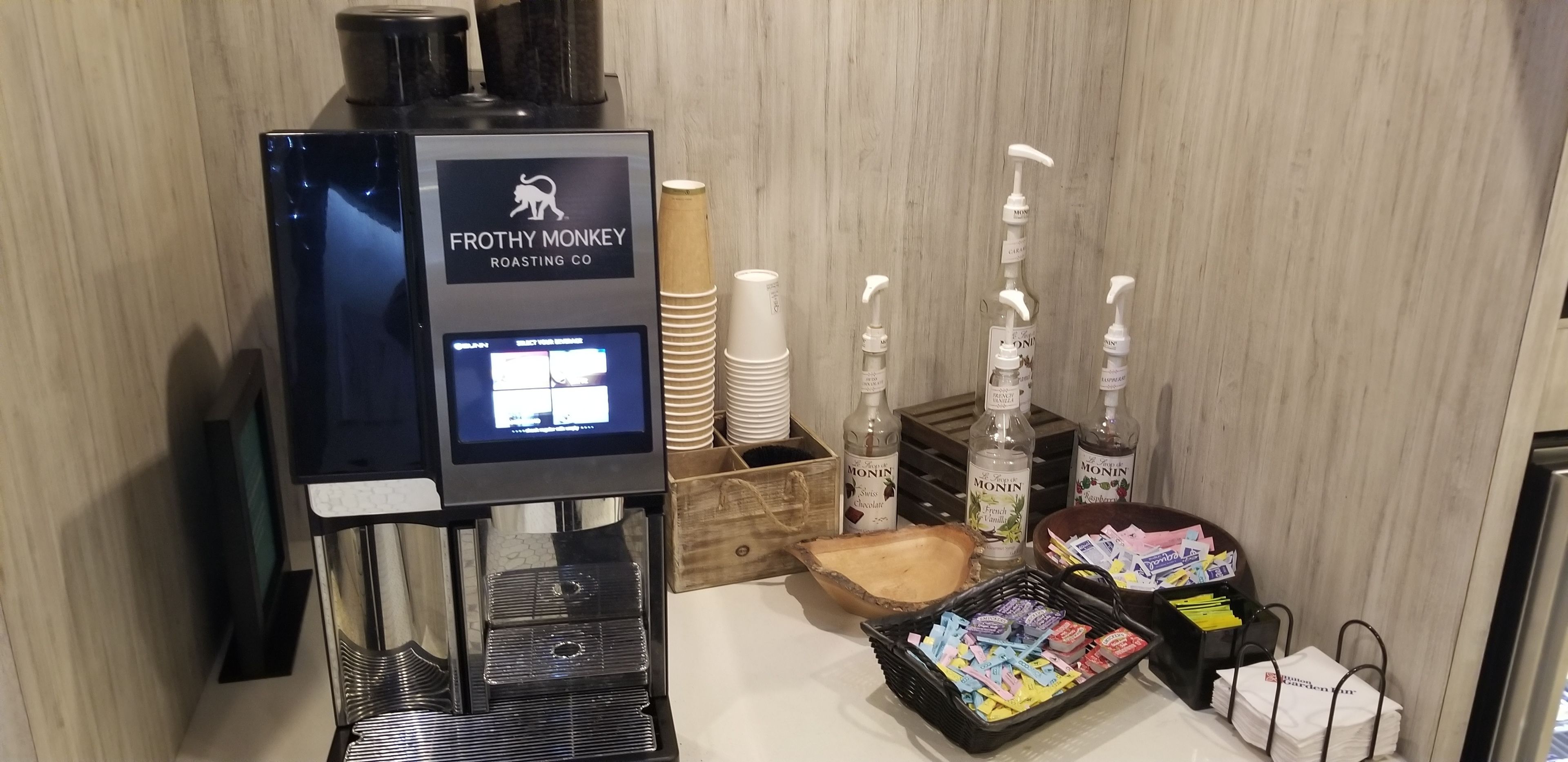 Un puesto de Frothy Monkey Roasting Co. con una pantalla de menú digital, vasos desechables, dispensadores de sirope Monin y una bandeja con diversos aperitivos.