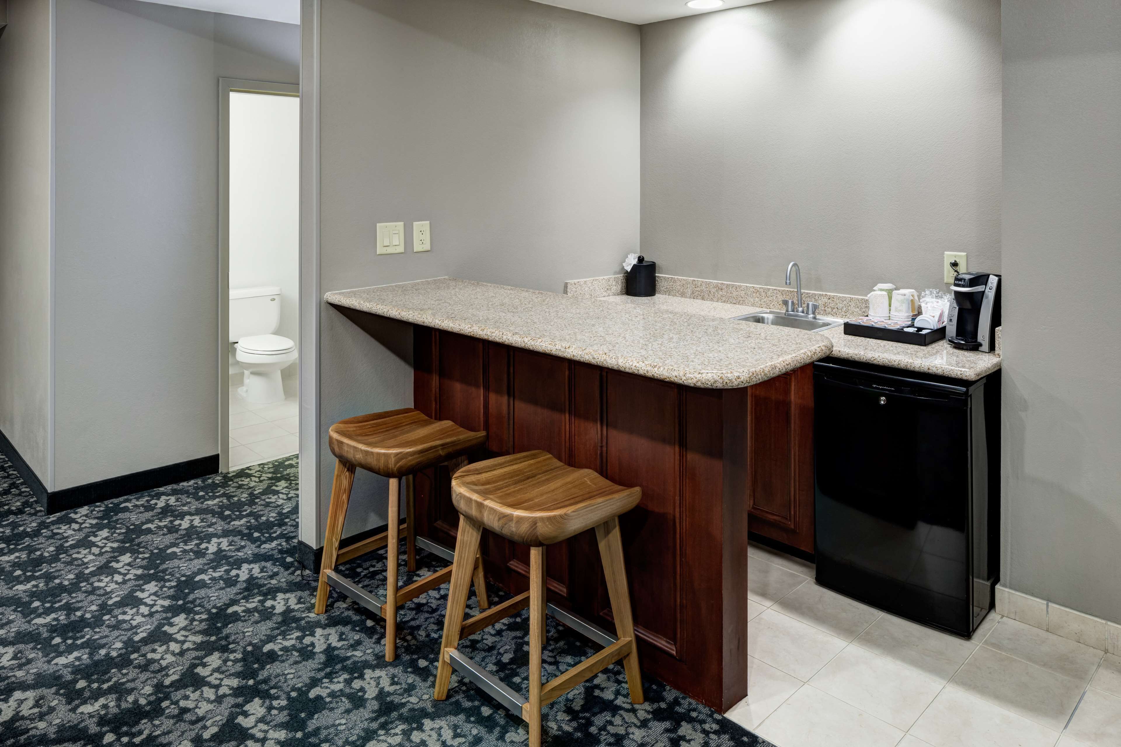 A compact kitchen with a granite countertop, two wooden stools, a dishwasher, and a sink.