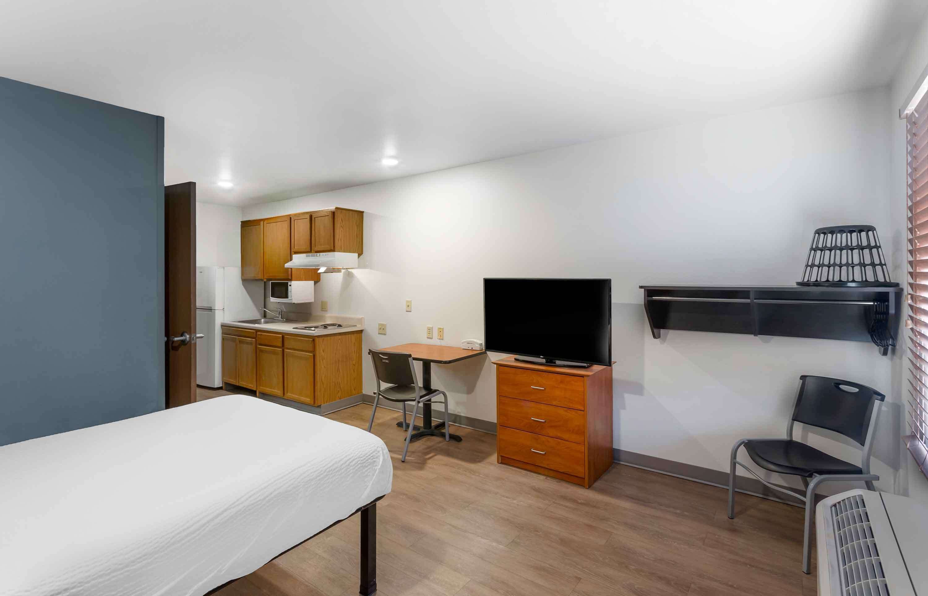 Habitación compacta con cama, escritorio y silla, televisión, kitchenette pequeña y estante con lámpara.