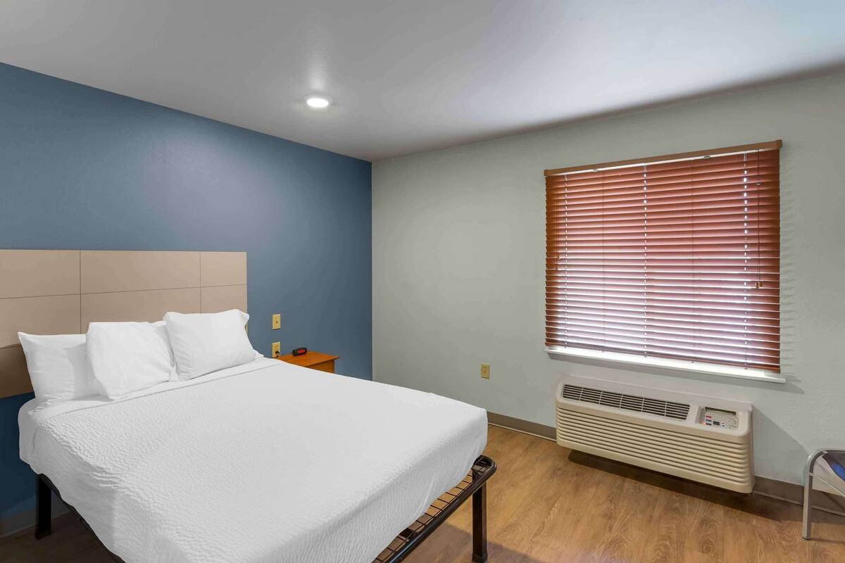 Un dormitorio con cama, mesita de noche, ventana con persianas y unidad de aire acondicionado.
