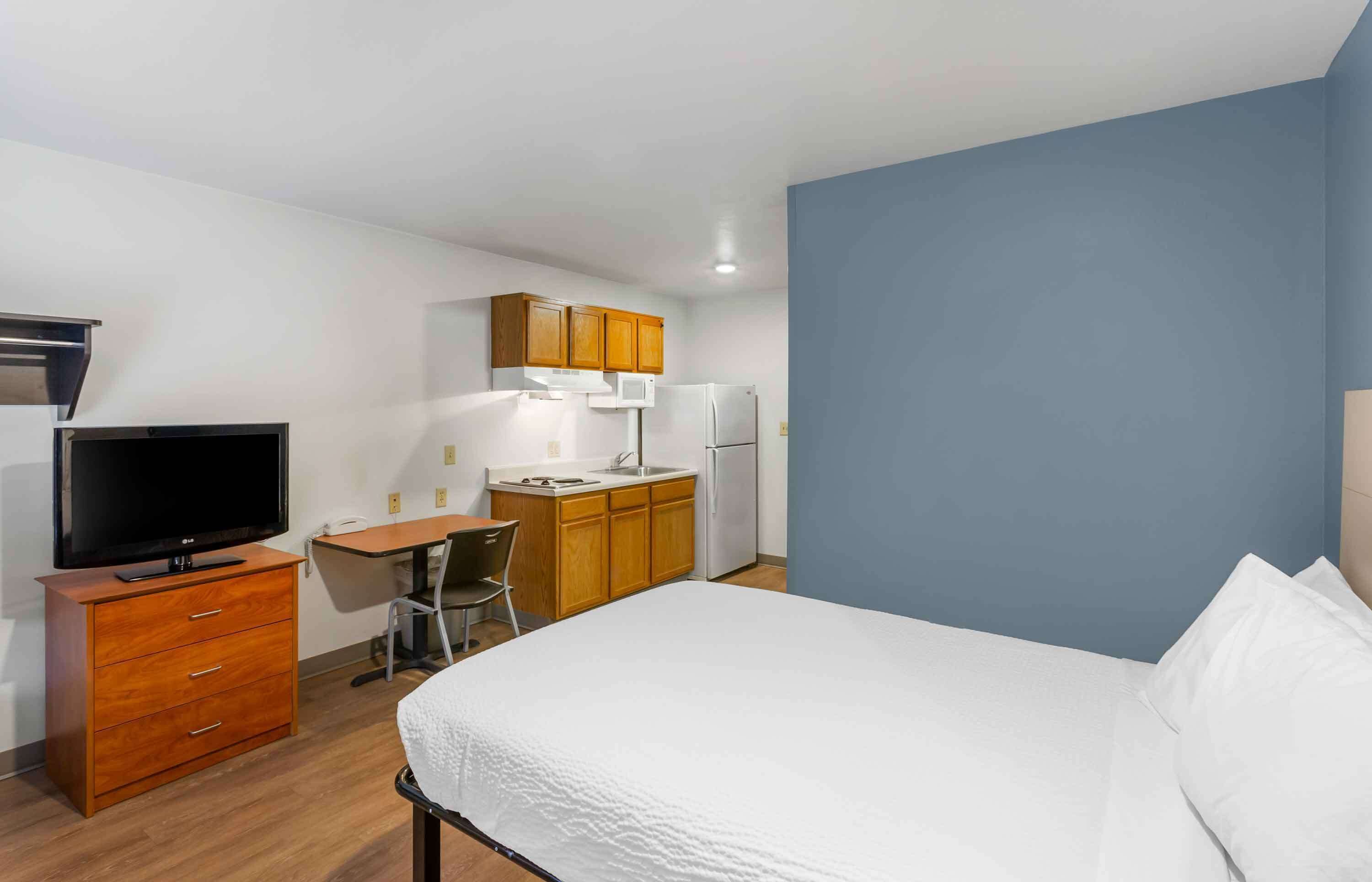 A compact hotel room with a bed, a desk, a television, and a kitchenette.