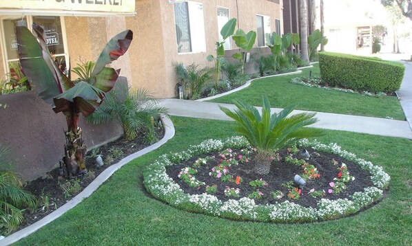 Un jardin bien entretenu, avec un parterre de fleurs circulaire, un sentier et un bâtiment sur lequel est apposée une enseigne indiquant « Hôtel ».