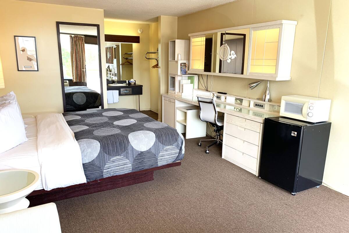 A hotel room with a bed, a desk, a chair, a small refrigerator, a microwave, a mirror, and a bathroom with a sink and towel rack.