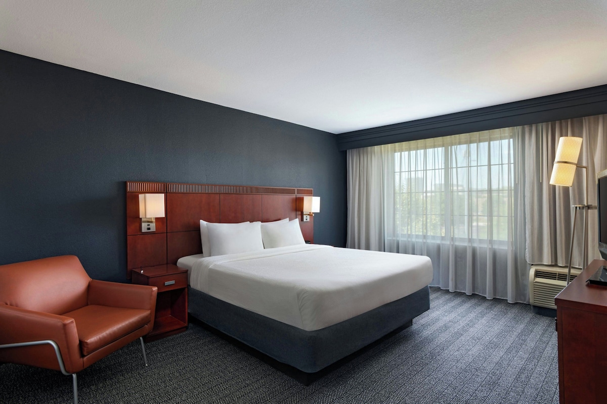 A hotel room with a large bed, a wooden headboard, a nightstand, a desk, a chair, and a window with curtains.