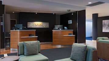 A modern reception area with two wooden reception desks, a seating area with green upholstered sofas, and a wall-mounted artwork.