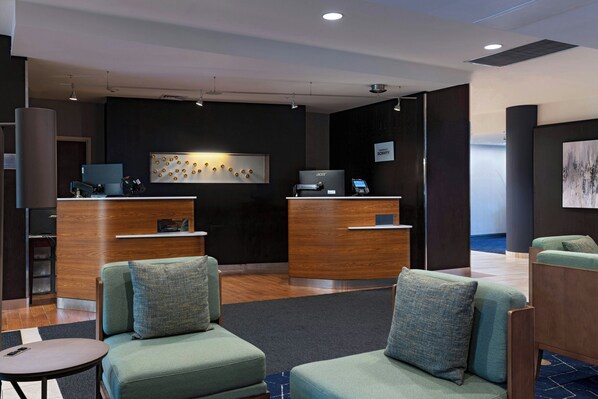 A modern reception area with two wooden reception desks, a seating area with green upholstered sofas, and a wall-mounted artwork.