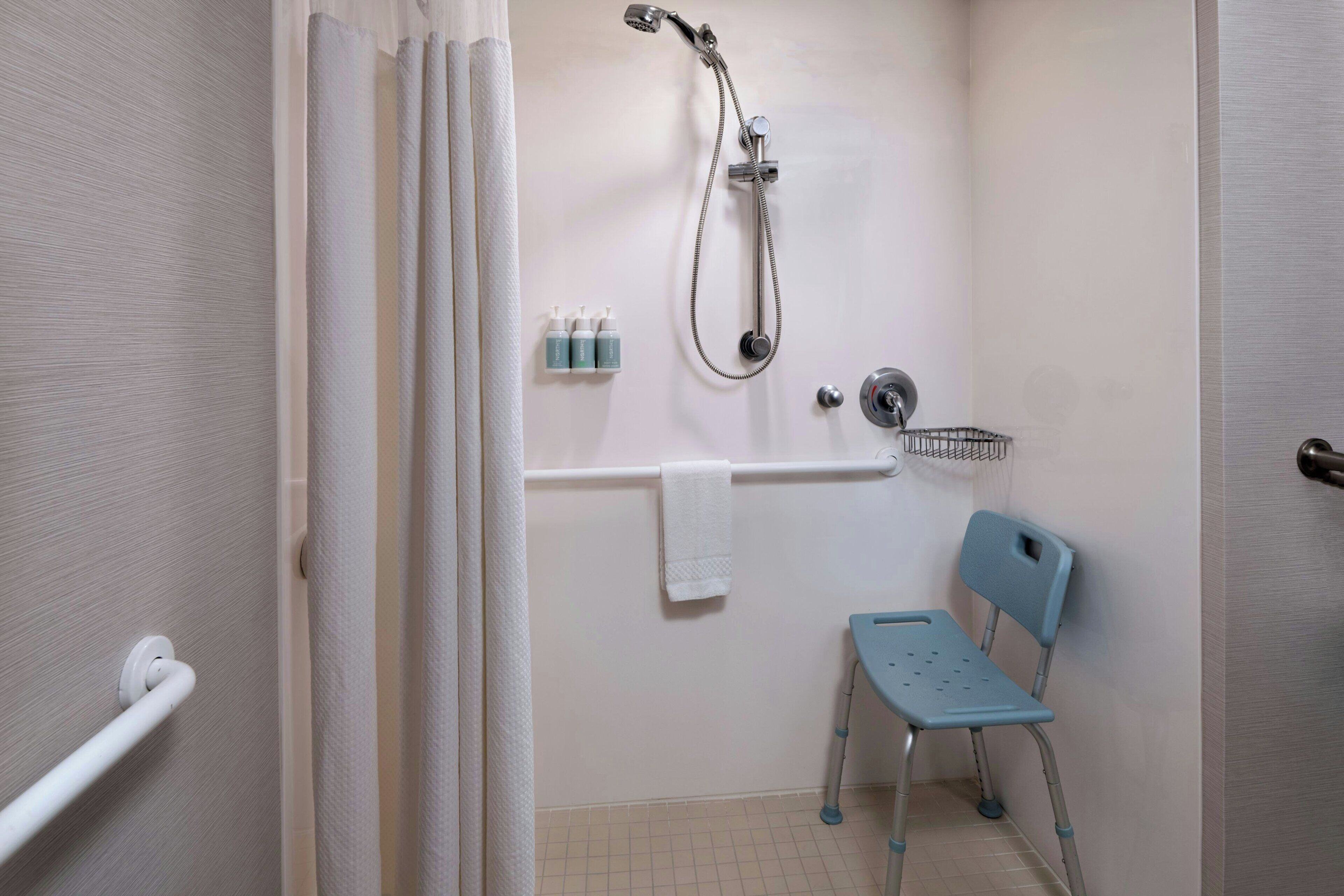 A bathroom with a shower, a grab bar, a wheelchair-accessible chair, and a towel rack.