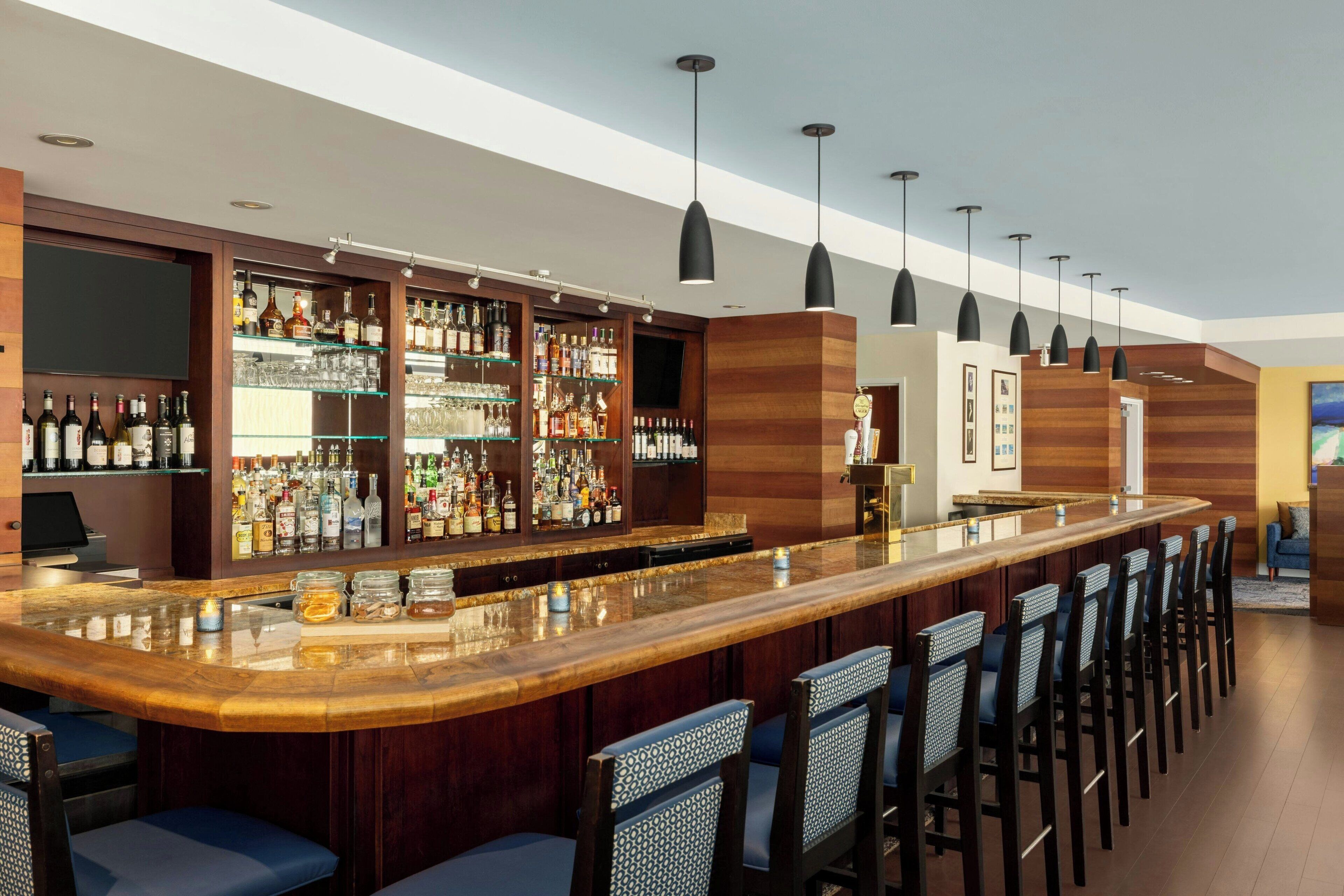 A bar area with a long counter, multiple shelves of liquor bottles, and several hanging pendant lights.