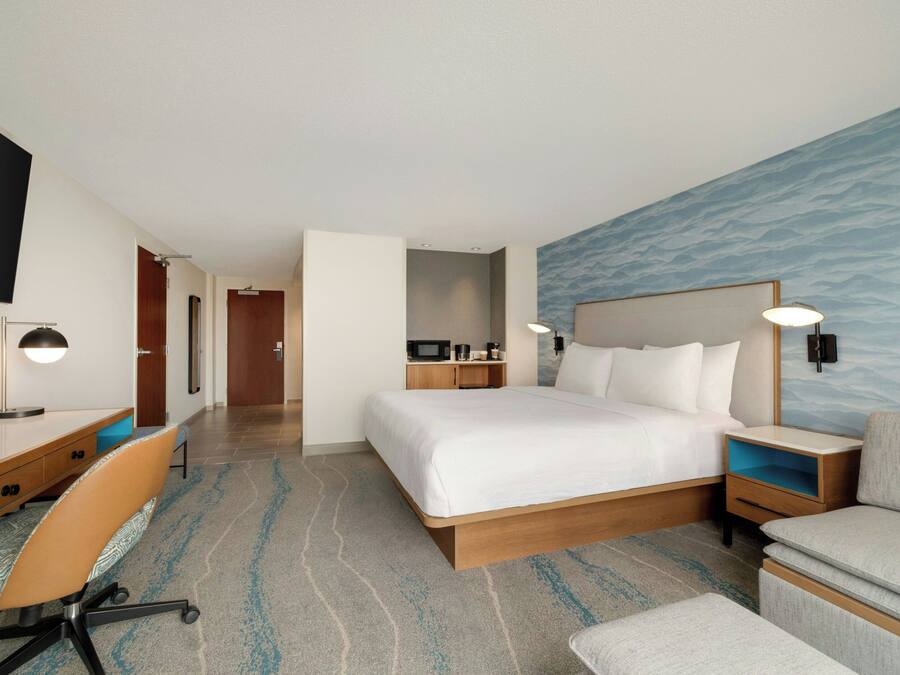 A hotel room with a bed, a desk with a chair, a TV, a microwave, a nightstand, and a wall-mounted lamp.