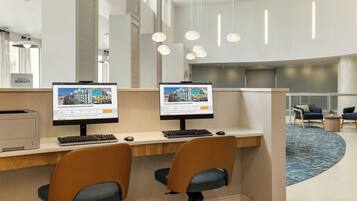 A modern hotel reception area with two computer workstations, a printer, and a seating area with chairs and a small table.