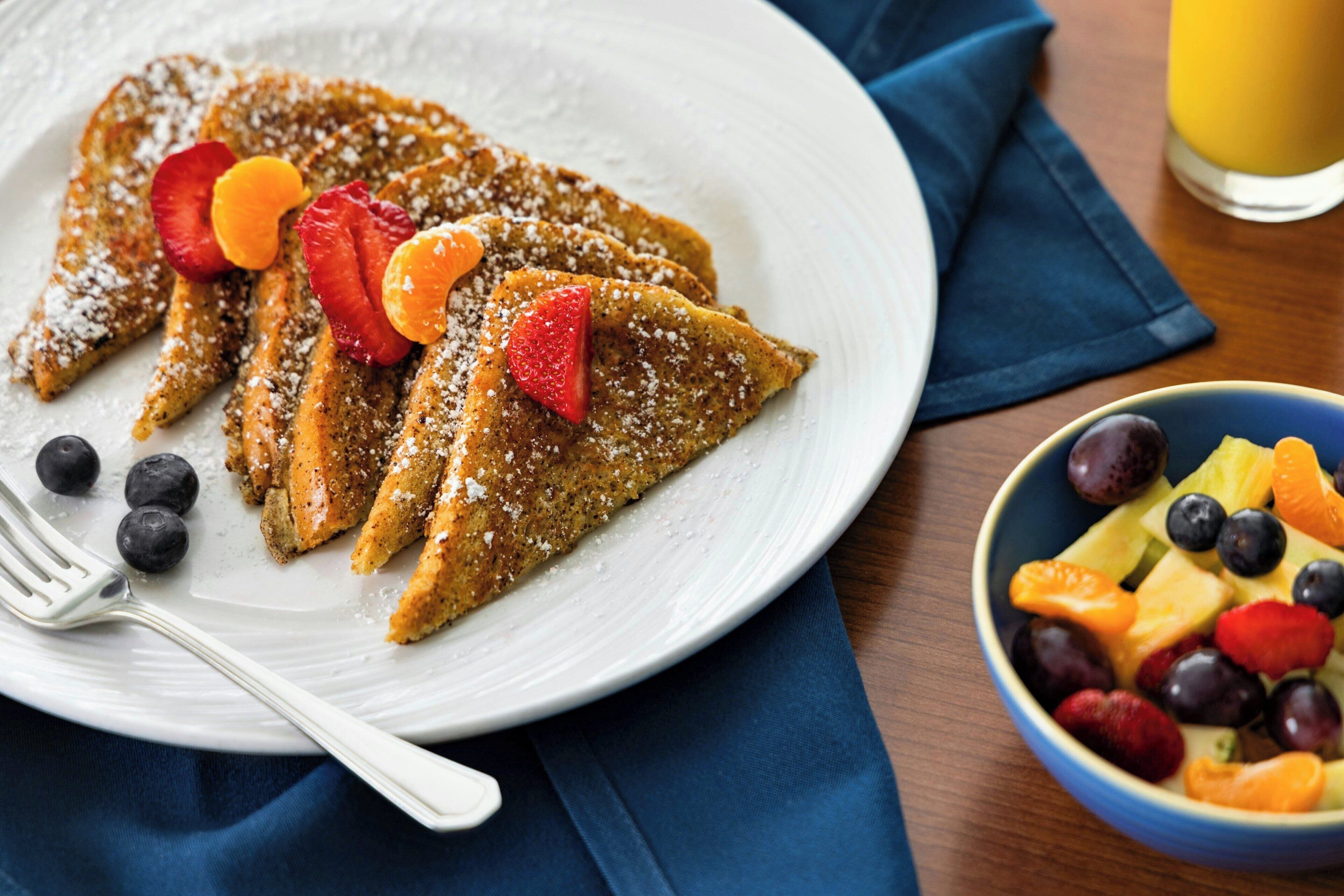 Ein Teller French Toast mit Obst und Puderzucker, eine Schüssel mit gemischtem Obst und ein Glas Orangensaft auf einem Holztisch.