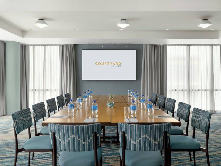 Sala de conferência com uma mesa comprida, cadeiras, uma tela exibindo 'COURTYARD BY MARRIOTT' e garrafas de água.
