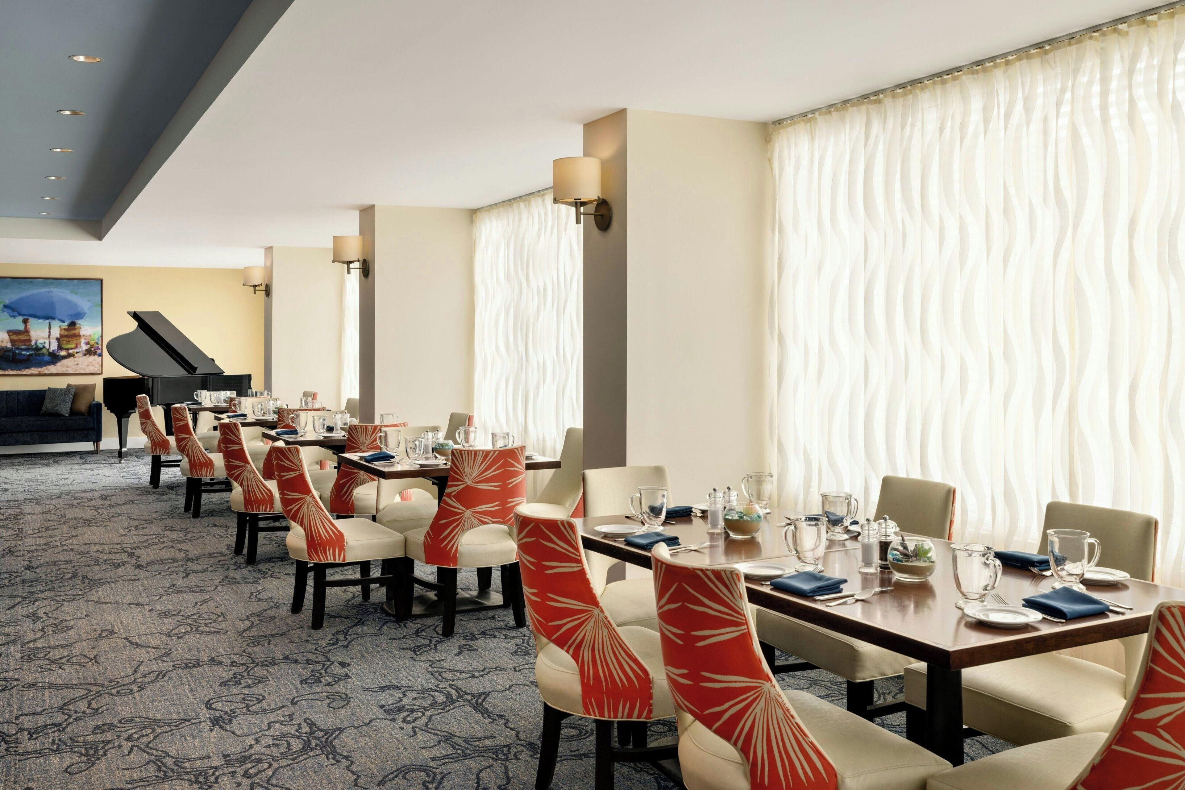 A dining area with tables set for four, chairs with patterned upholstery, and a grand piano in the background.