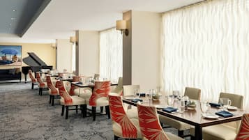 A dining area with tables set for four, chairs with patterned upholstery, and a grand piano in the background.