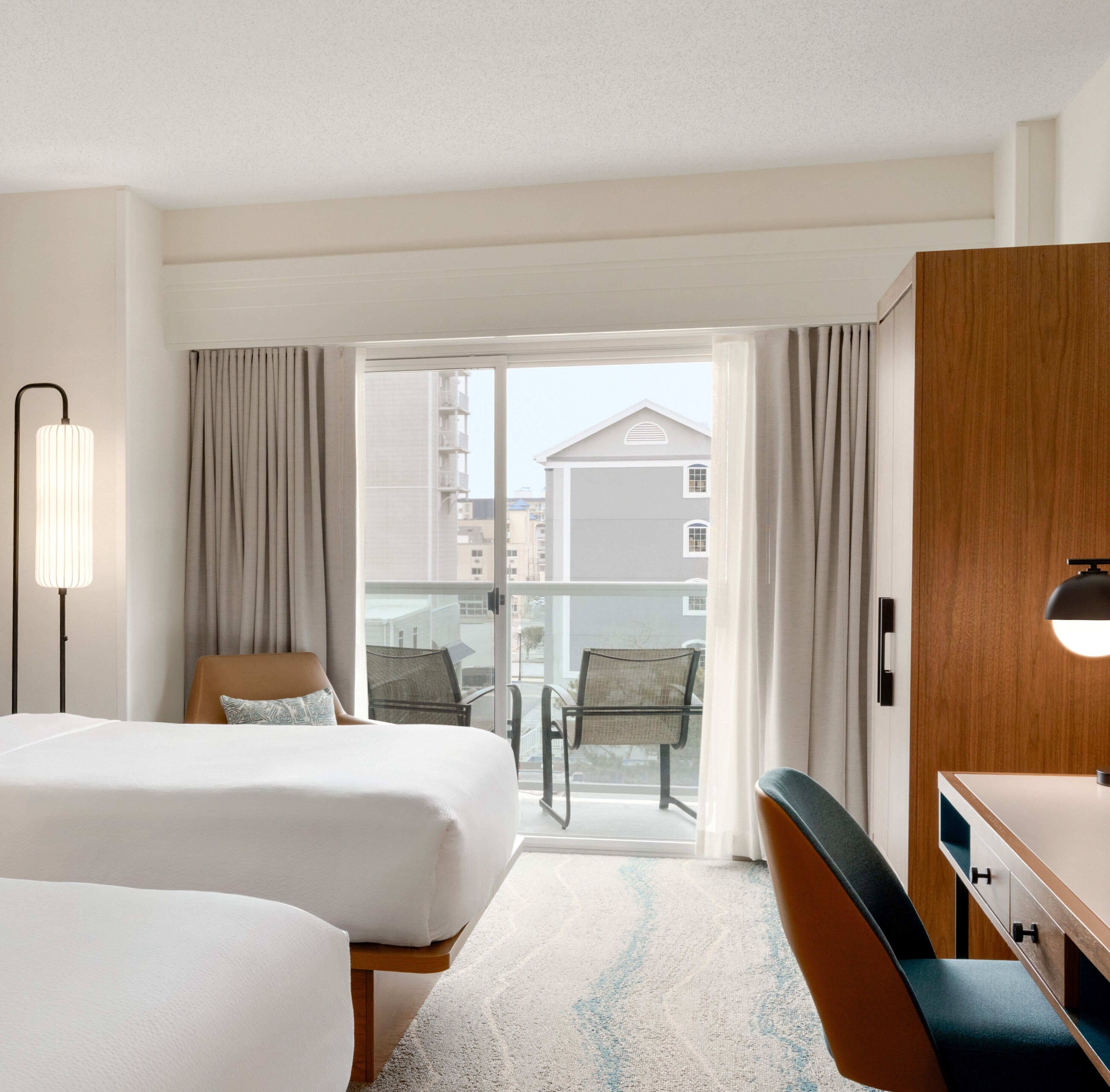 A hotel room with two beds, a desk, a chair, a lamp, and a view of buildings through a glass door.