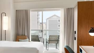 A hotel room with two beds, a desk, a chair, a lamp, and a view of buildings through a glass door.