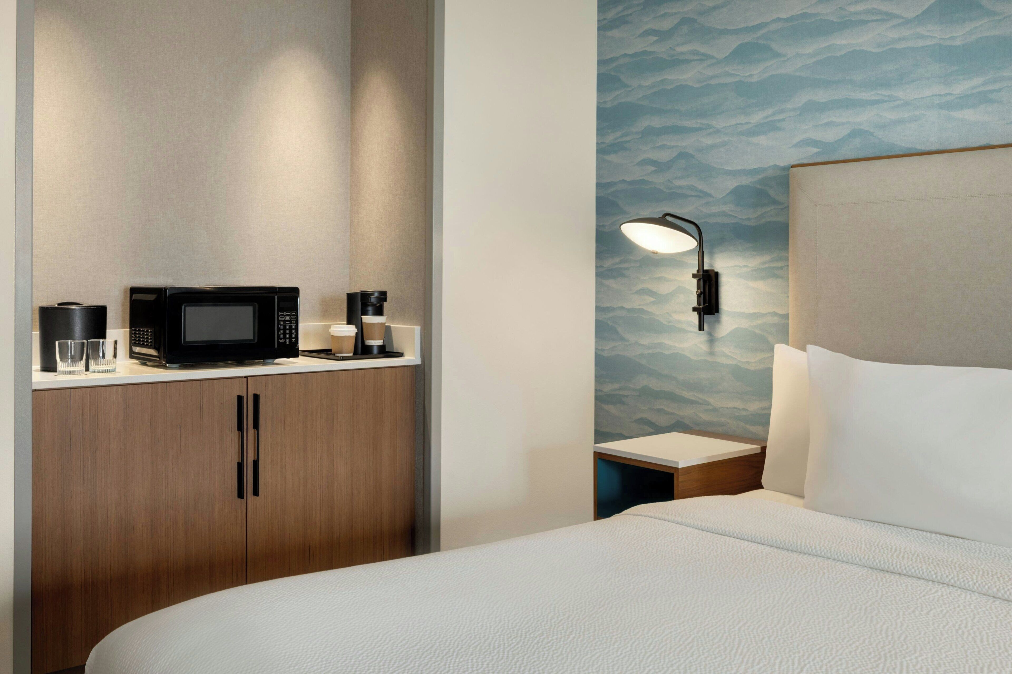 Une chambre d’hôtel équipée d’un lit, d’un four à micro-ondes, d’une machine à café et d’une table de chevet avec une lampe.