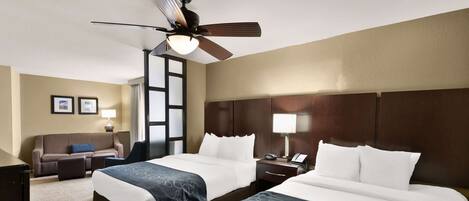 Ein Hotelzimmer mit zwei Betten, einem Deckenventilator, einem Schreibtisch mit Lampe, einem Sofa und einem großen Fenster.