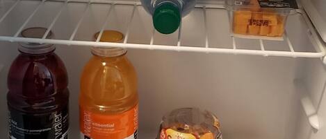 A refrigerator containing various food and drink items including soda cans, a water bottle, a vitamin drink, and snacks.