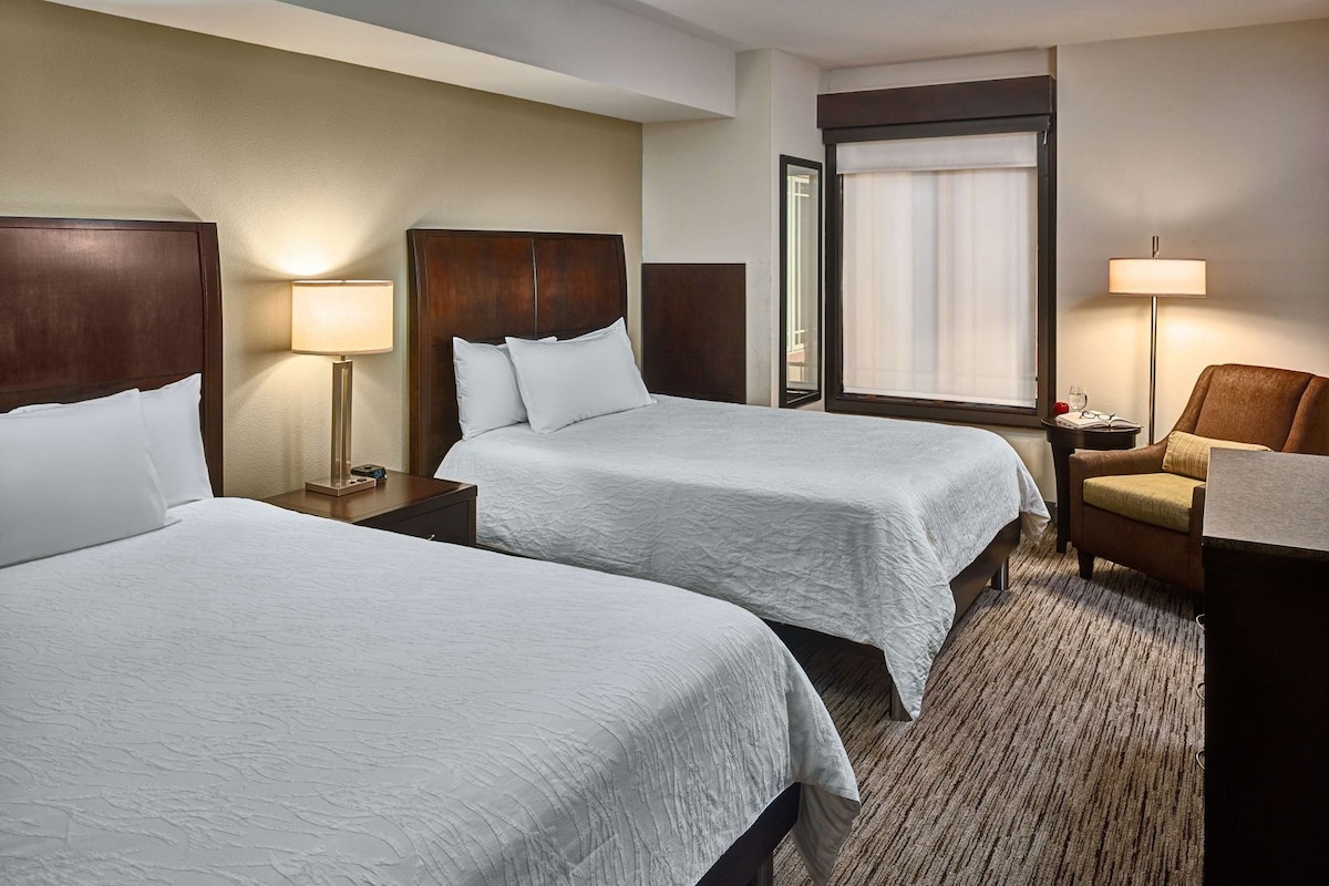 A hotel room with two beds, a chair, a lamp, a nightstand, and a window with curtains.
