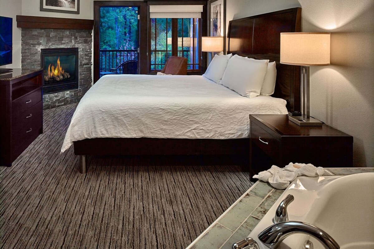 A hotel room with a large bed, a fireplace, a view of trees, a desk with a lamp, and a bathtub.