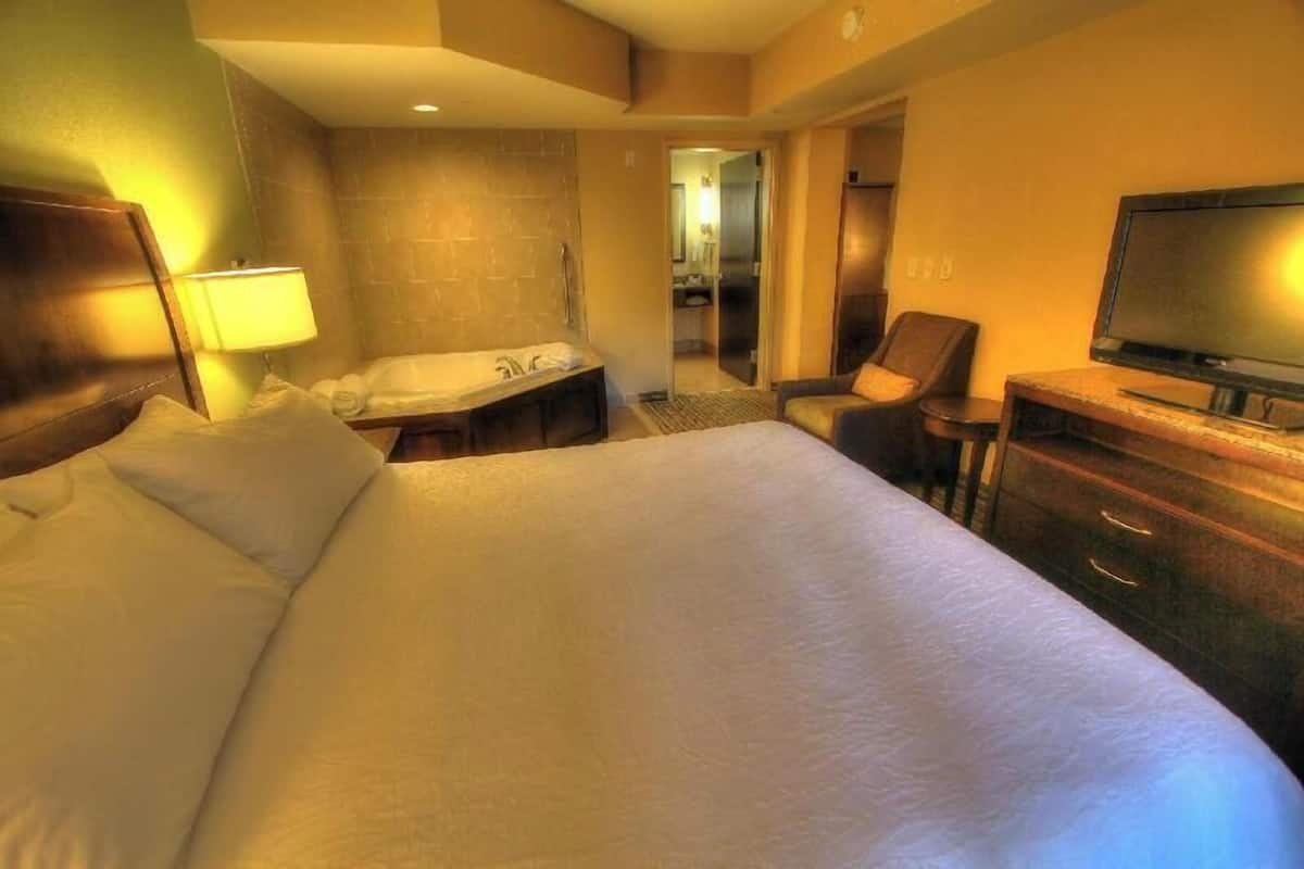 Ein Hotelzimmer mit einem großen Bett, einem Schreibtisch, einem Fernseher, einem Badezimmer inklusive Waschbecken und Spiegel sowie einem Sessel.