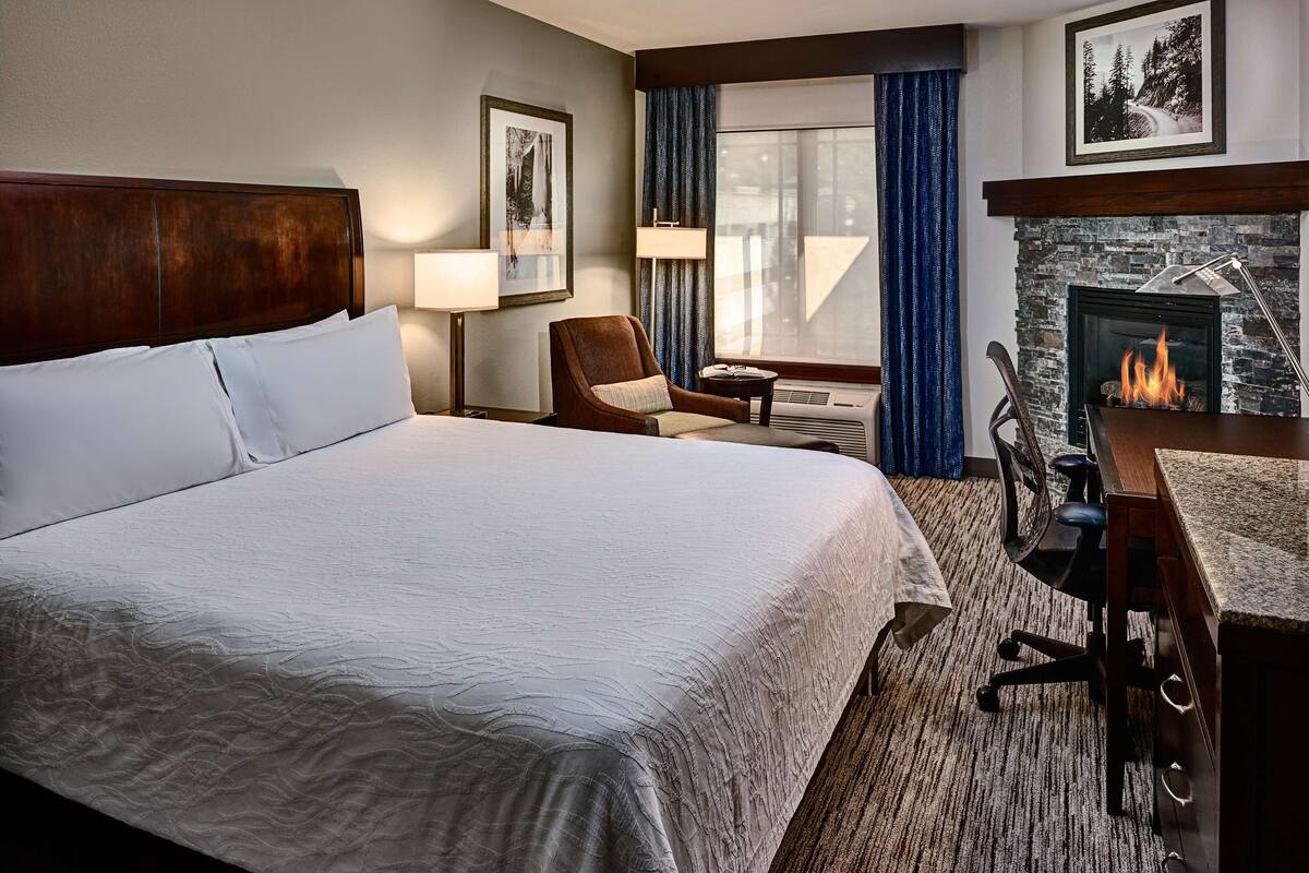 A hotel room with a large bed, a desk, a chair, a fireplace, and a window with curtains.