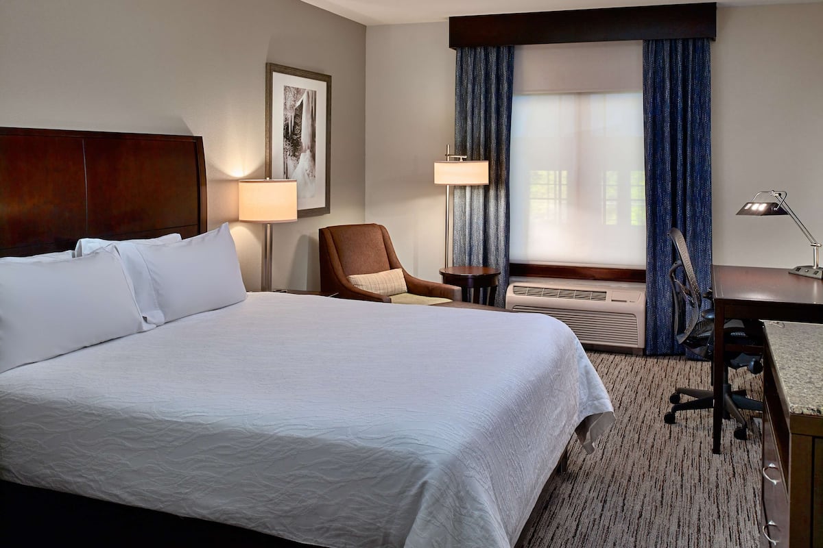 A hotel room with a large bed, a desk with a chair, a lamp, a picture on the wall, and a window with curtains.