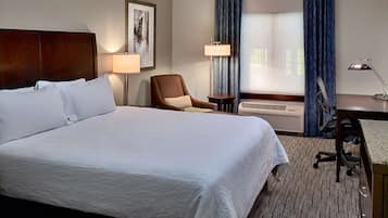 Habitación de hotel con una cama grande, un escritorio con una silla, una lámpara, un cuadro en la pared y una ventana con cortinas.