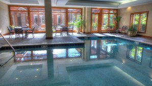 An indoor swimming pool with tiled flooring, surrounded by wooden-framed windows and potted plants.