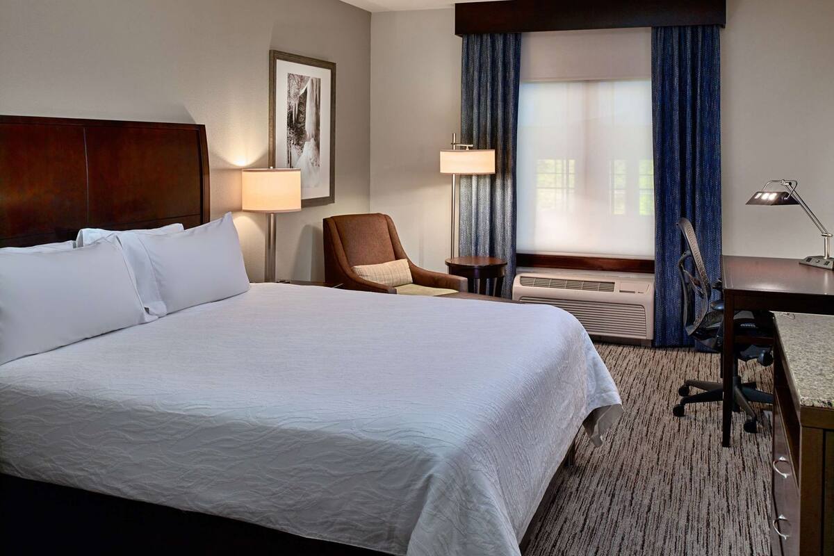 A hotel room with a large bed, a desk, a chair, a lamp, a picture on the wall, and a window with curtains.