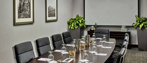 A conference room with a long table, black leather chairs, framed pictures on the wall, and potted plants.