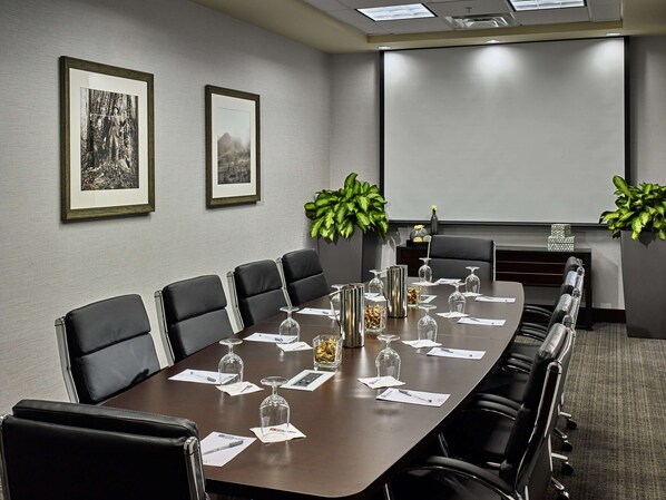 Un salón de conferencias con una larga mesa, sillas de cuero negro, cuadros en la pared y plantas en macetas.